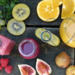A top view of assorted fresh fruits including kiwi, oranges, figs, raspberries, grapes, and various colorful fruit juices in bottles and a jug on a rustic wooden table.